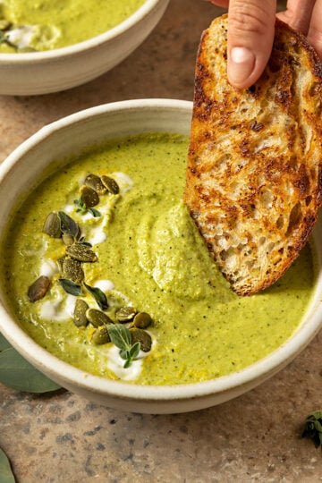 hand dipping toasted bread into a bowl of broccoli soup