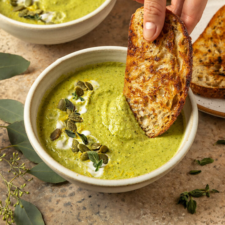 hand dipping toasted bread into a bowl of broccoli soup