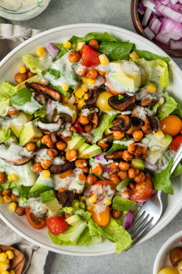 bowl of cobb salad with lettuce, chickpeas, mushroom bacon, red onion, tomato, corn, and yogurt ranch dressing surrounded by sides.
