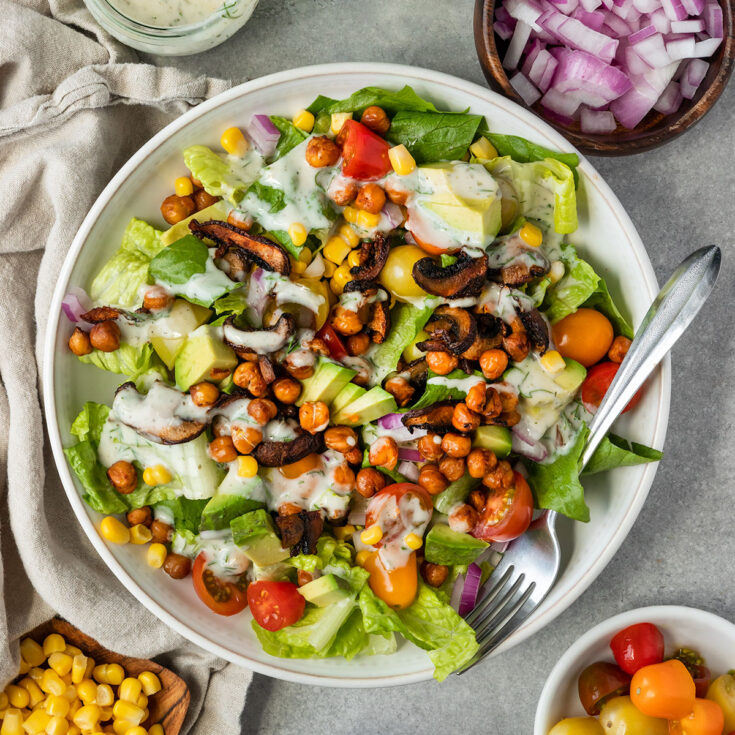 bowl of cobb salad with lettuce, chickpeas, mushroom bacon, red onion, tomato, corn, and yogurt ranch dressing surrounded by sides.