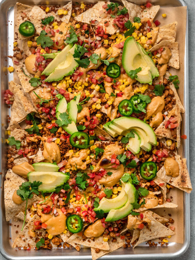 tray of nachos with vegan queso, pico de gallo, avocado slices, jalapeno slices, corn, and tofu crumbles.
