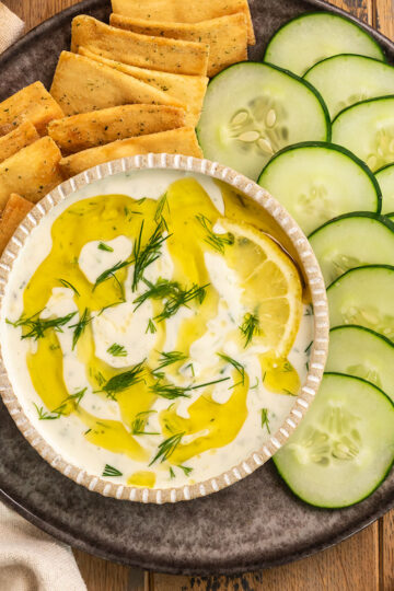 bowl of tzatziki on plate with crackers, cucumber slices, and lemons.