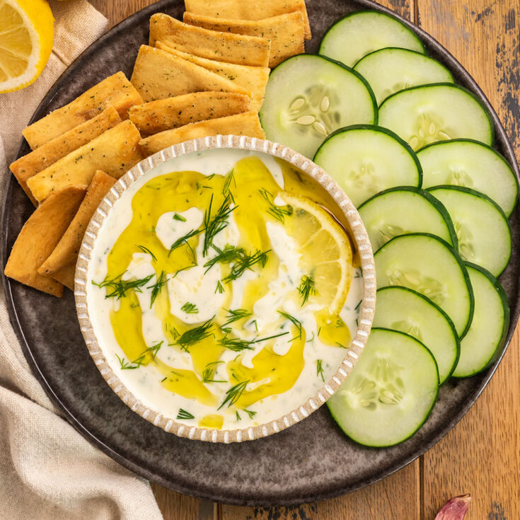 bowl of tzatziki on plate with crackers, cucumber slices, and lemons.