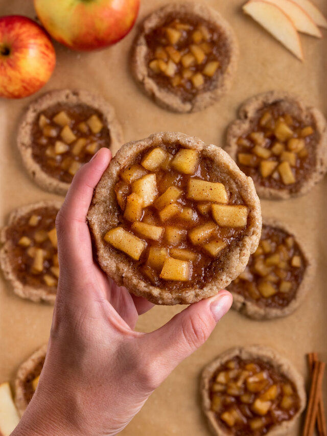 hand holding apple pie filled cookie over tray of apple pie cookies.