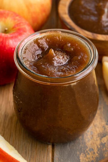 glass container of apple butter surrounded by fresh apples.