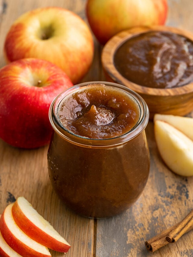 glass container of apple butter surrounded by fresh apples.