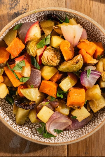 bowl of roasted potatoes, sweet potatoes, apples, red onion, brussel sprouts, and carrot topped with parsley.
