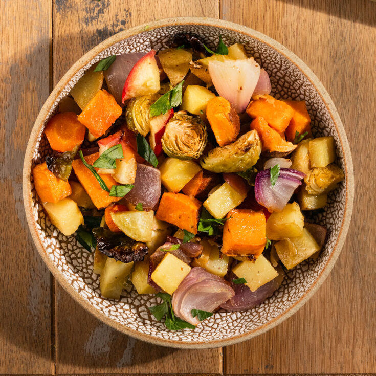 bowl of roasted potatoes, sweet potatoes, apples, red onion, brussel sprouts, and carrot topped with parsley.