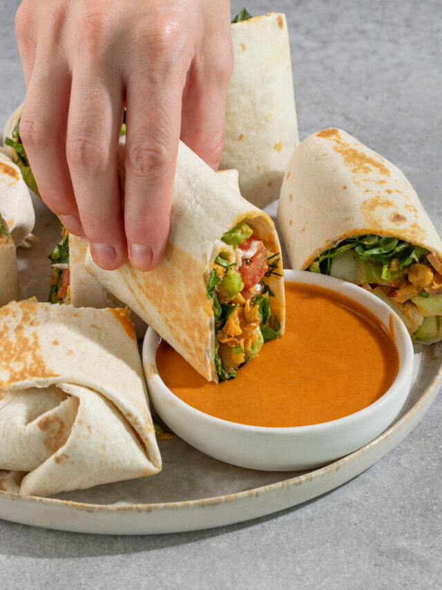 dipping a wrap filled with chickpeas lettuce avocado and tomato into a cup of buffalo sauce.