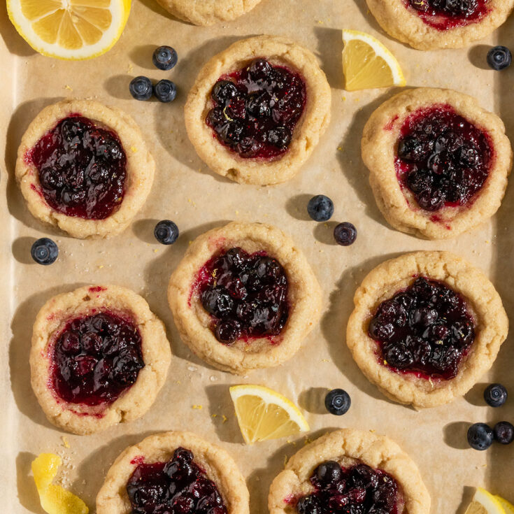 tray of baked lemon cookies filled with blueberry pie filling surrounded by lemon slices and fresh blueberries.