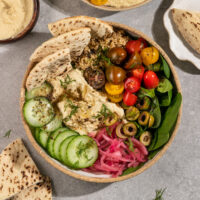 a bowl filled with spinach, quinoa, hummus, cucumber, pickled red onions, olives, cherry tomatoes, pita, fresh dill, and dressing.