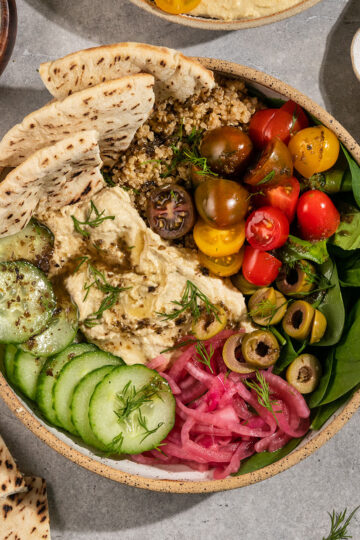 a bowl filled with spinach, quinoa, hummus, cucumber, pickled red onions, olives, cherry tomatoes, pita, fresh dill, and dressing.