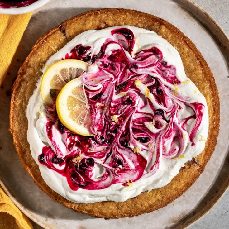 round cake topped with swirls of whipped cream, blueberry jam, lemon slices, and lemon zest.