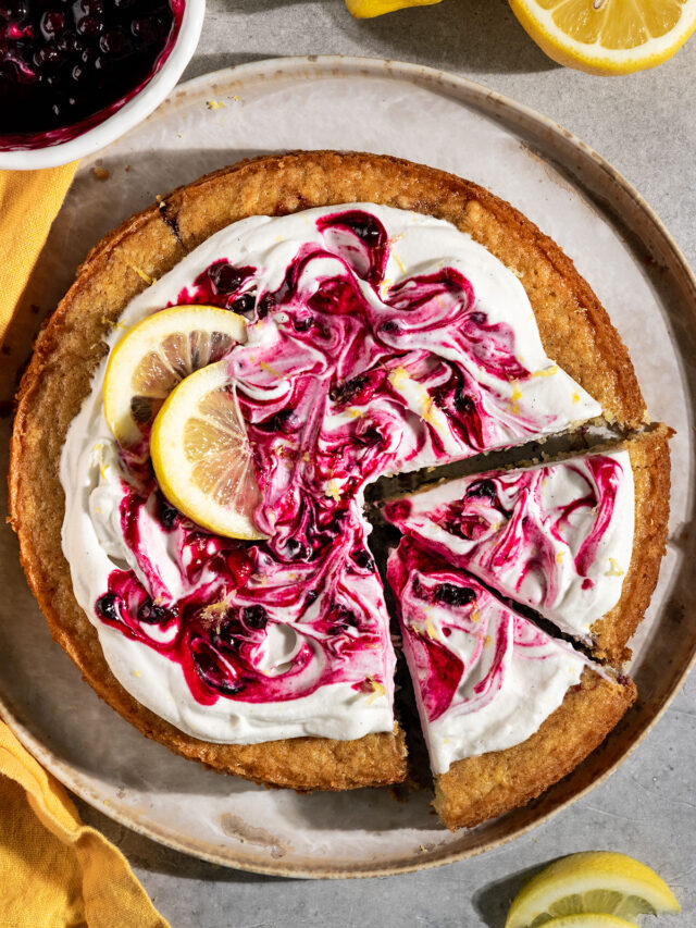 round cake topped with swirls of whipped cream, blueberry jam, and lemon slices with two slices cut out.