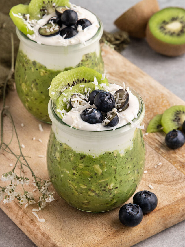 two jars of matcha overnight oats topped with yogurt blueberries kiwi and dried coconut.