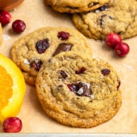 two cranberry orange chocolate chip cookies layered on top of eachother on tray with more cookies, fresh orange slices, and fresh cranberries.