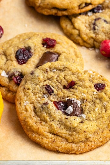 two cranberry orange chocolate chip cookies layered on top of eachother on tray with more cookies, fresh orange slices, and fresh cranberries.