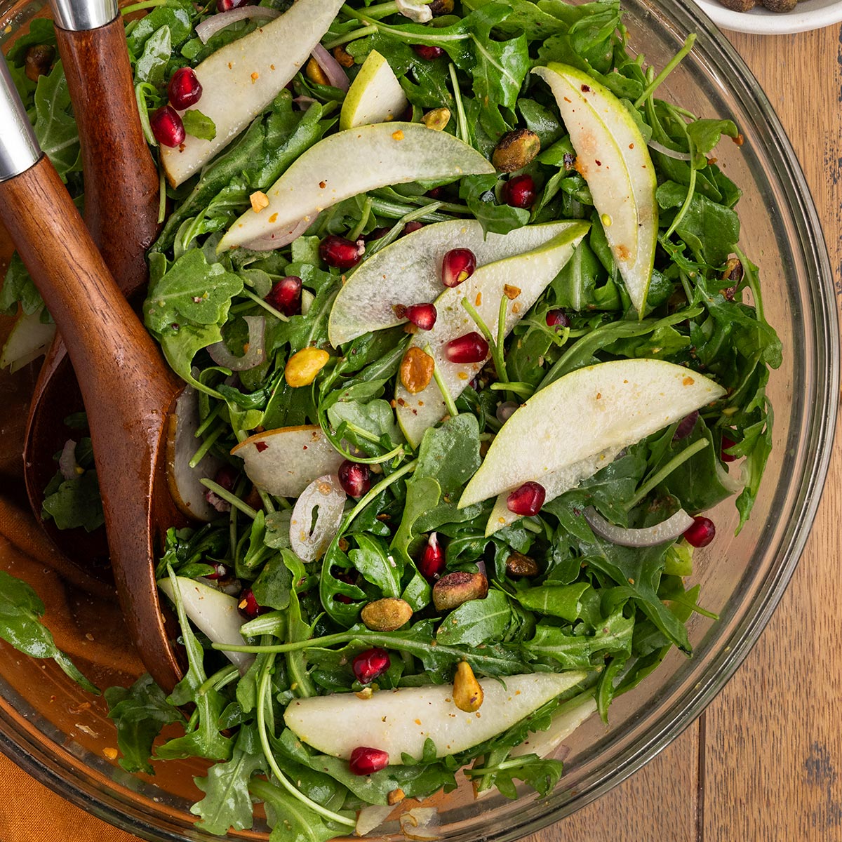 bowl of salad with arugula, pears, pistachios, pomegranate seeds, dressing and two wooden salad spoons.