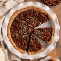 sliced pecan pie in a pie dish alongside a slice of pecan pie, pecan halves, and whipped cream.