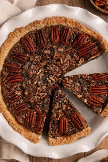 sliced pecan pie in a pie dish alongside a slice of pecan pie, pecan halves, and whipped cream.