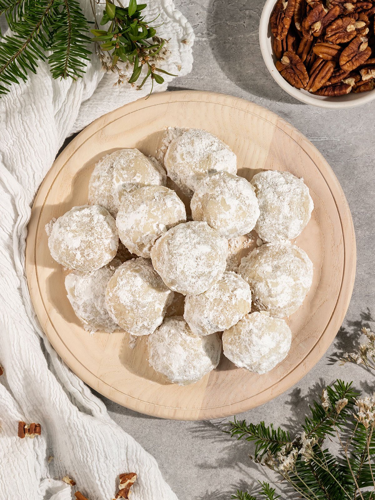 plate of powdered sugar coated snowball cookies besides bowl of pecans and greenery.