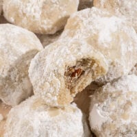 plate of powdered sugar snowball cookies with top cookie missing a bite and showing inside full of pecans.