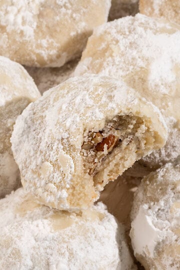 plate of powdered sugar snowball cookies with top cookie missing a bite and showing inside full of pecans.