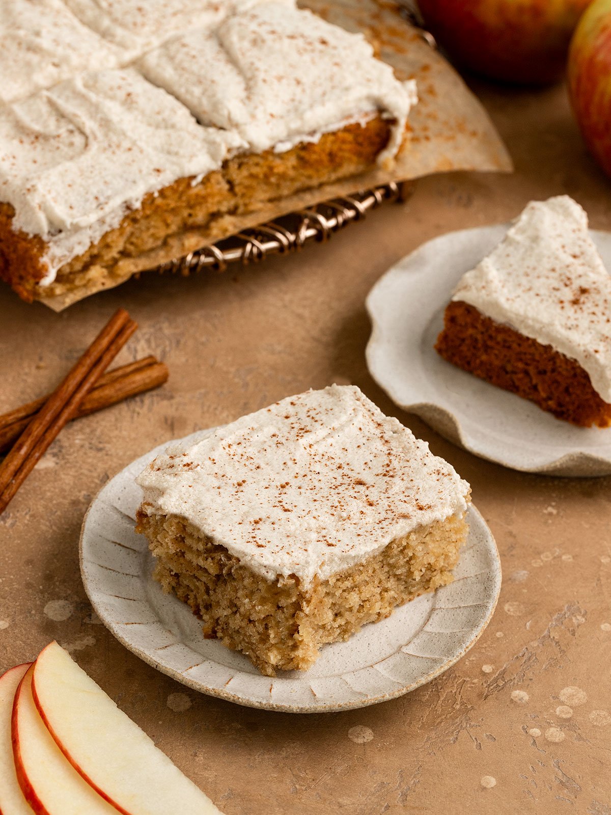 two slices of apple cake with frosting and cinnamon sprinkled on top on plates alongside cinnamon sticks and fresh apple slices.