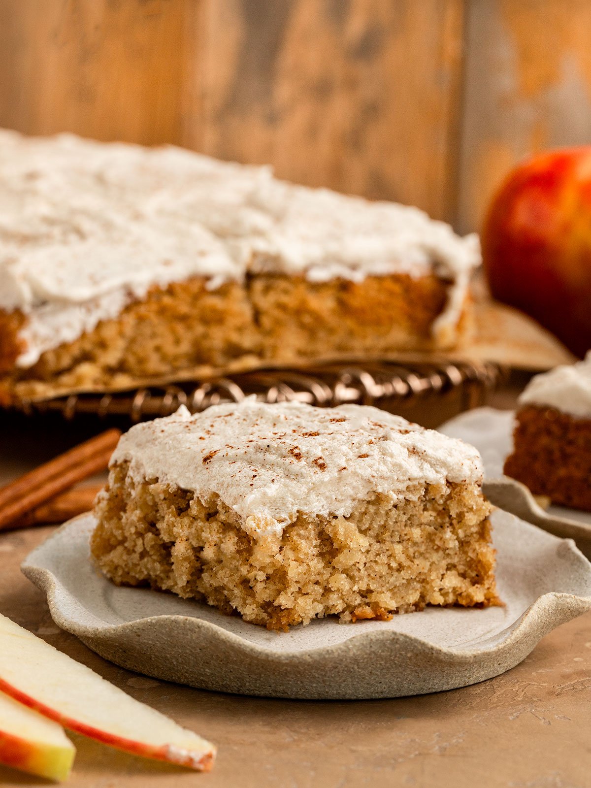 slice of cinnamon frosted  apple cake in front of whole cake on a metal cake stand.