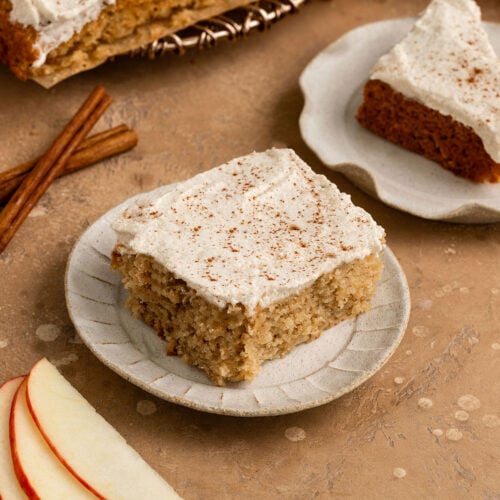 two slices of apple cake with frosting and cinnamon sprinkled on top on plates alongside cinnamon sticks and fresh apple slices.