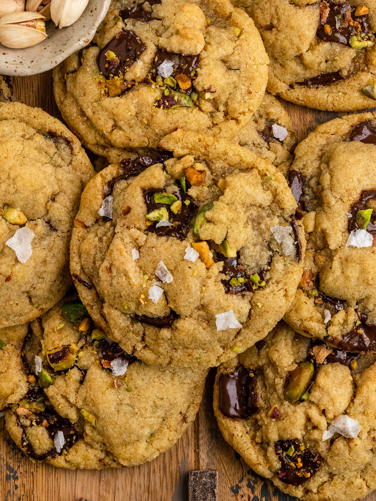 several olive oil chocolate chunk and pistachio cookies layered on top of each other aside bowl of pistachios.
