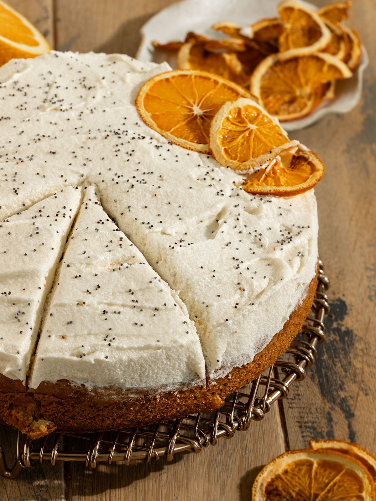 sliced orange poppyseed cake with cream cheese frosting and dried orange slices on a drying rack.