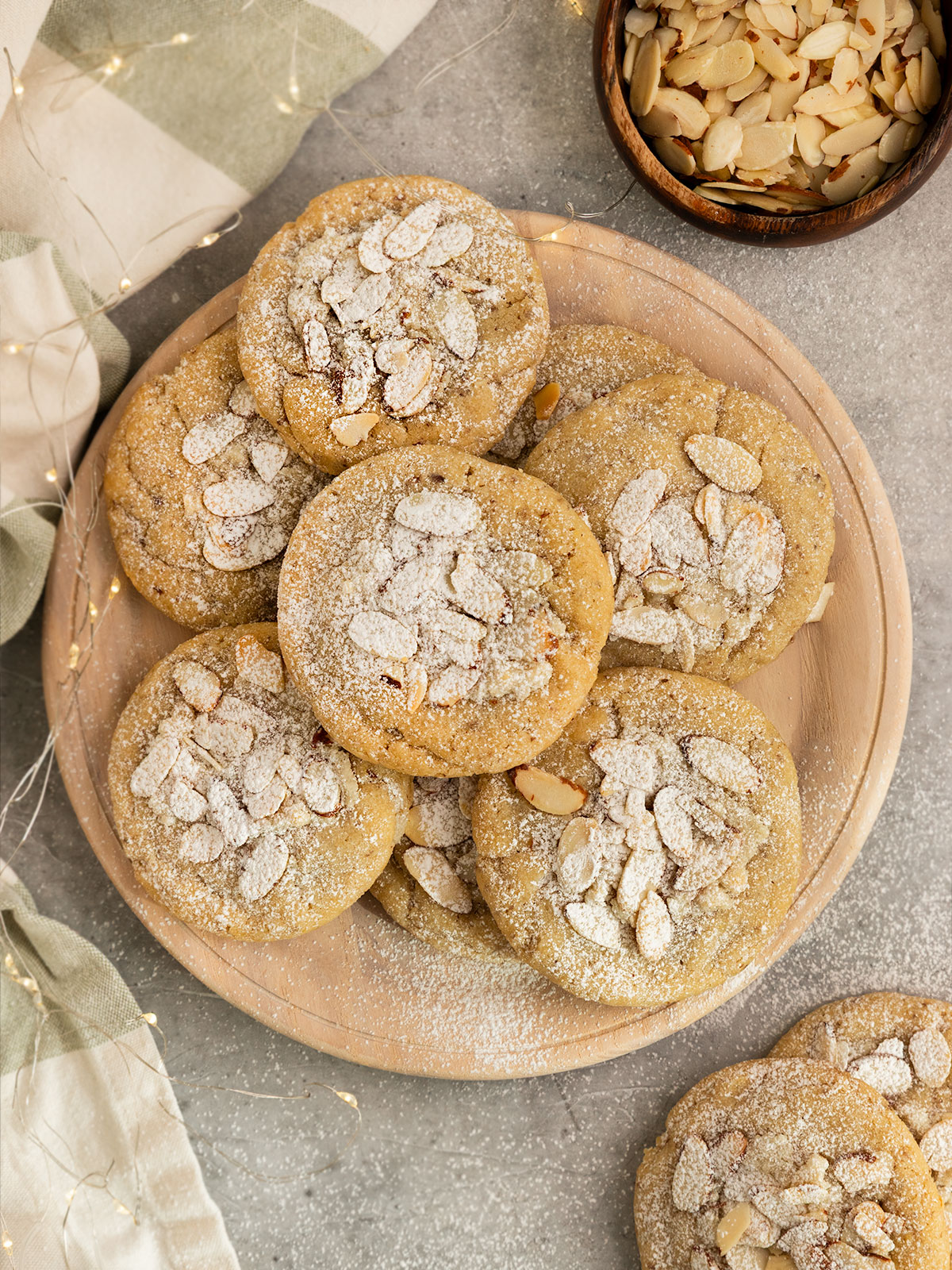 wooden plate of almond cookies topped with sliced almonds and powdered sugar aside a bowl of sliced almonds and lights.
