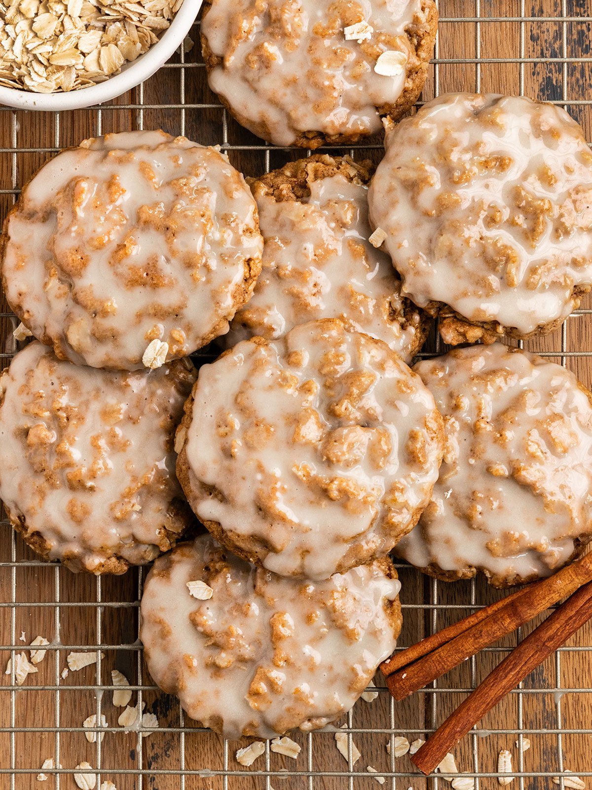 stack of iced oatmeal cookies with a bowl of oats and cinnamon sticks on a wire rack.