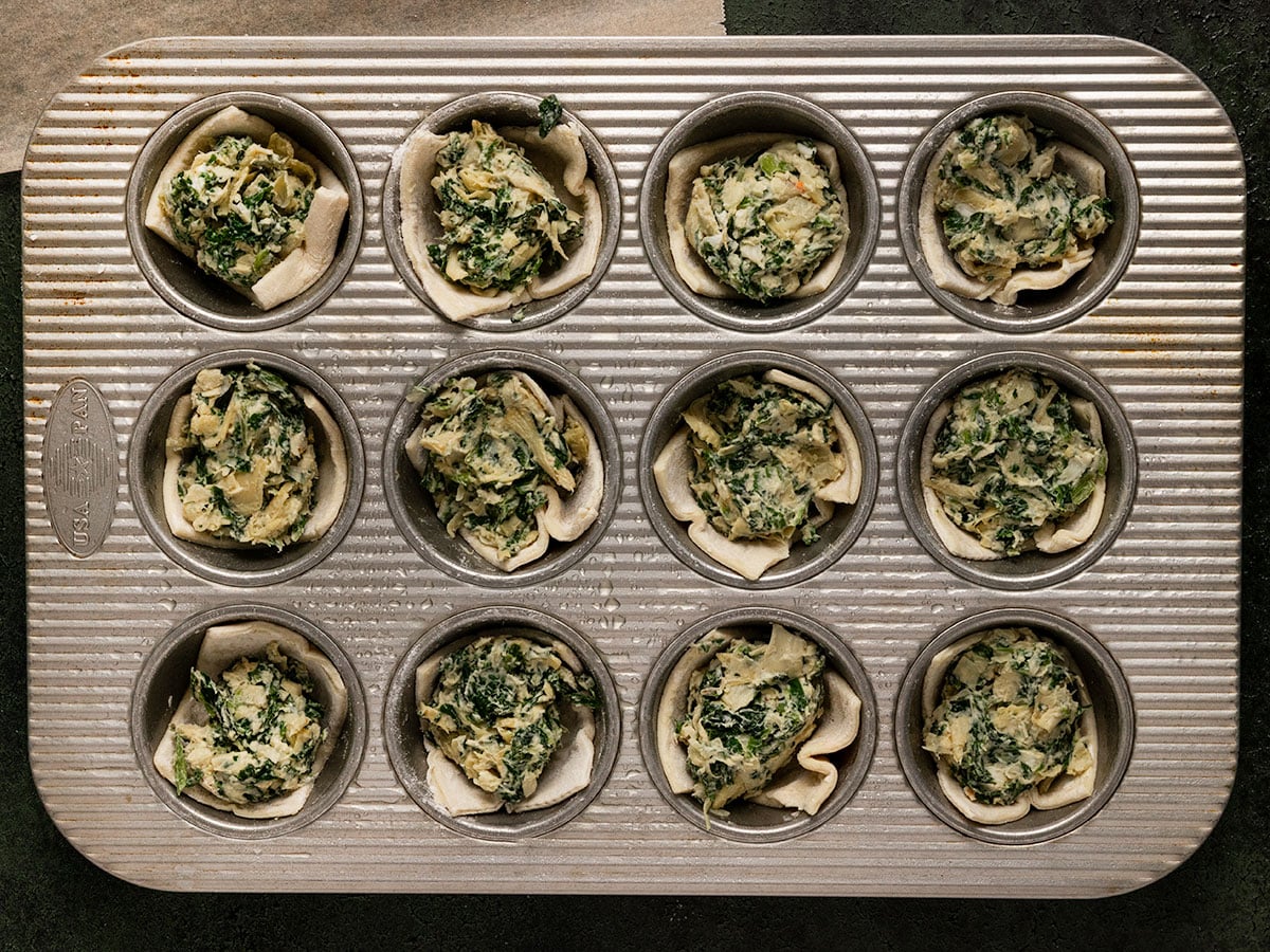 unbaked puff pastry slices in muffin tin filed with spinach artichoke filling.