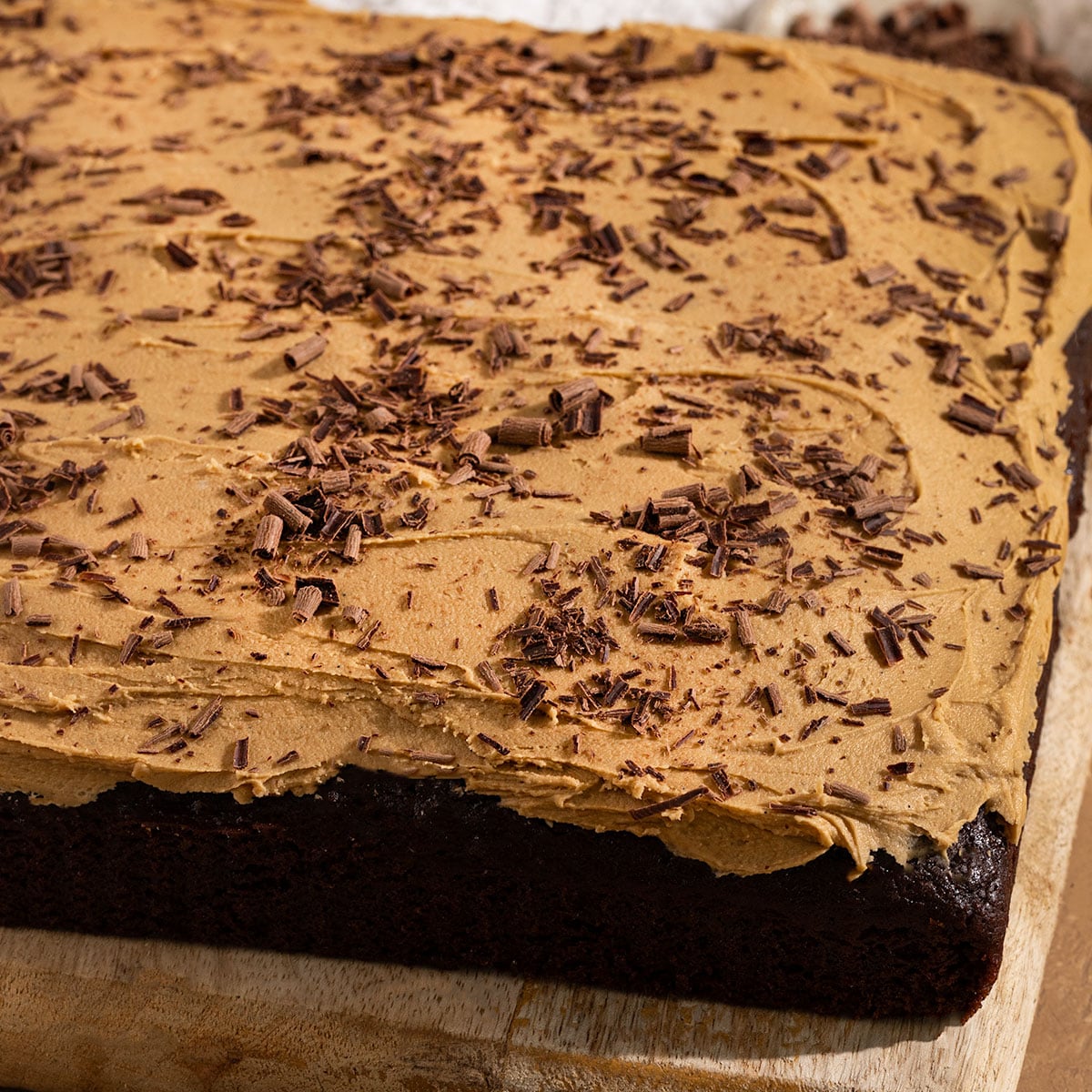 square shaped chocolate cake with brown sugar frosting and chocolate shavings on top of a wooden board.