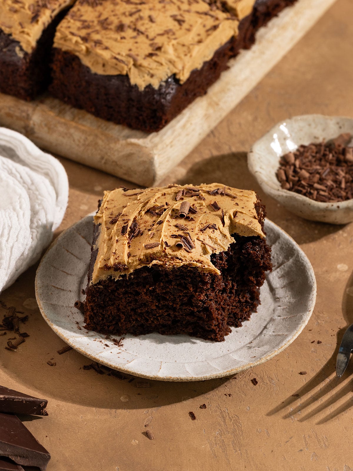 slice of chocolate cake with brown frosting and chocolate shavings with a bite taken out a fork on the side.