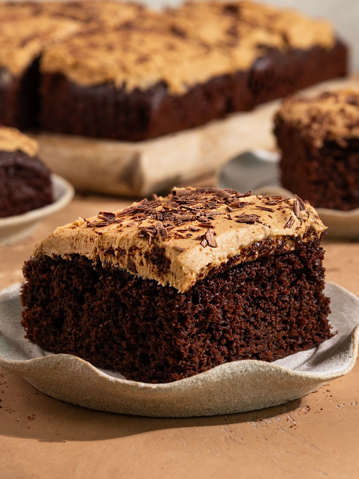 a slice of chocolate cake with brown frosting and chocolate shavings on a plate with the rest of the cake in the background.