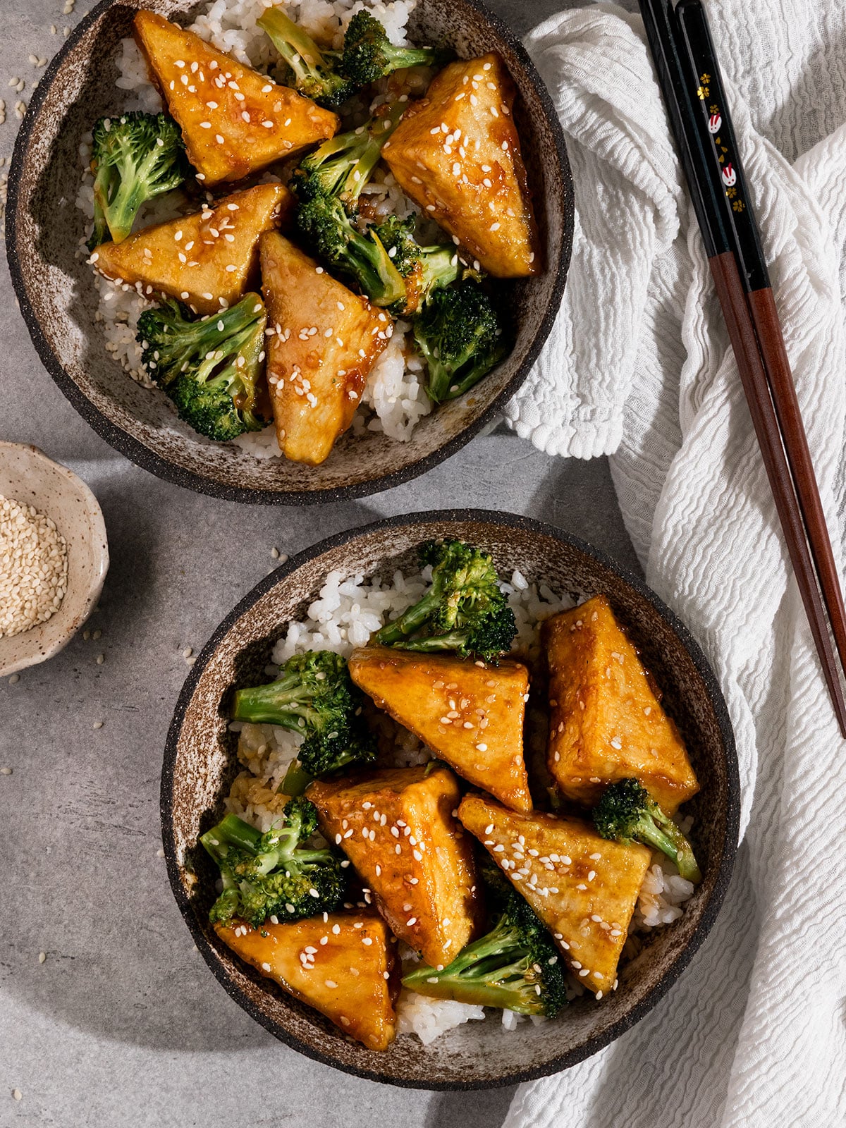 two bowls filled with white rice, broccoli, and tofu in a brown teriyaki sauce with chopsticks on the side.