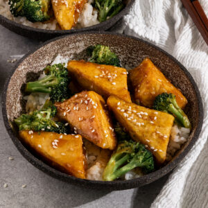 bowl filled with white rice, broccoli, and tofu in a brown teriyaki sauce with chopsticks on the side.