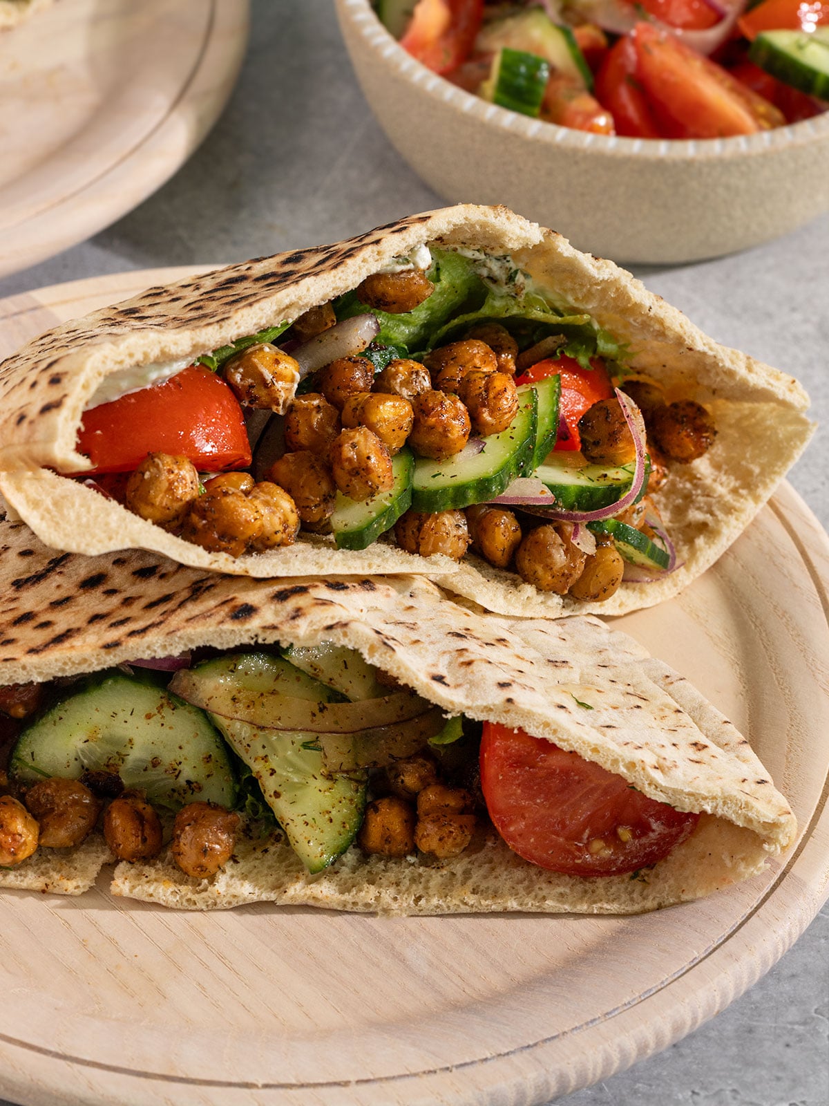 two pita pockets on a plate stuffed with crispy chickpeas, lettuce, tomato, onion, and cucumber.