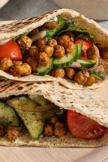 two pita pockets on a plate stuffed with crispy chickpeas, lettuce, tomato, onion, and cucumber.