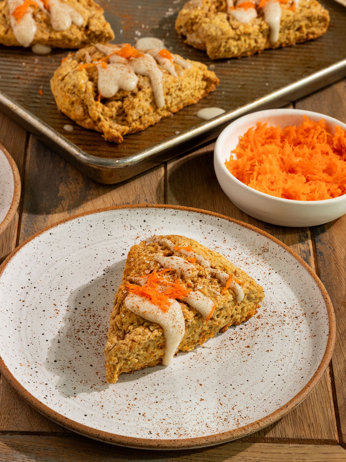 carrot cake scone on a plate topped with cream cheese glaze, cinnamon, and grated carrot alongside a tray of baked scones and bowl of more grated carrots.