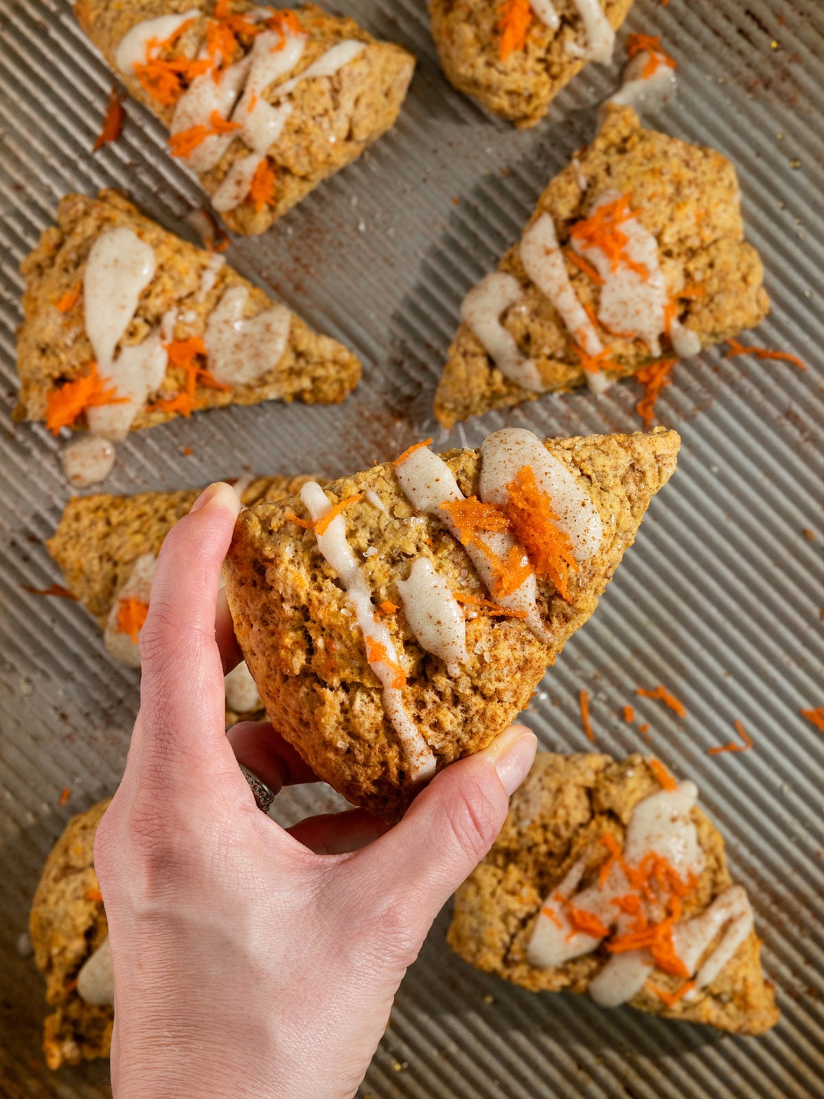 hand holding carrot scone over a tray of scones topped with cream cheese icing, ground cinnamon, and grated carrots.