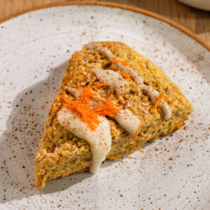triangle carrot cake scone on a plate topped with cream cheese glaze, ground cinnamon, and grated carrots.
