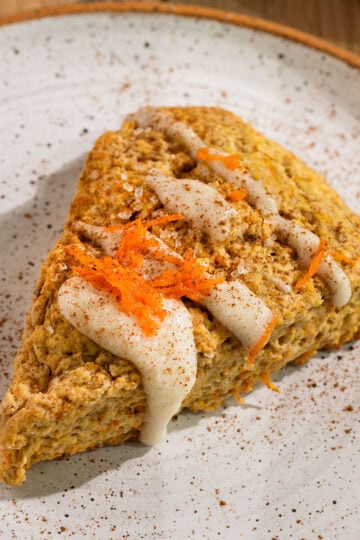 triangle carrot cake scone on a plate topped with cream cheese glaze, ground cinnamon, and grated carrots.