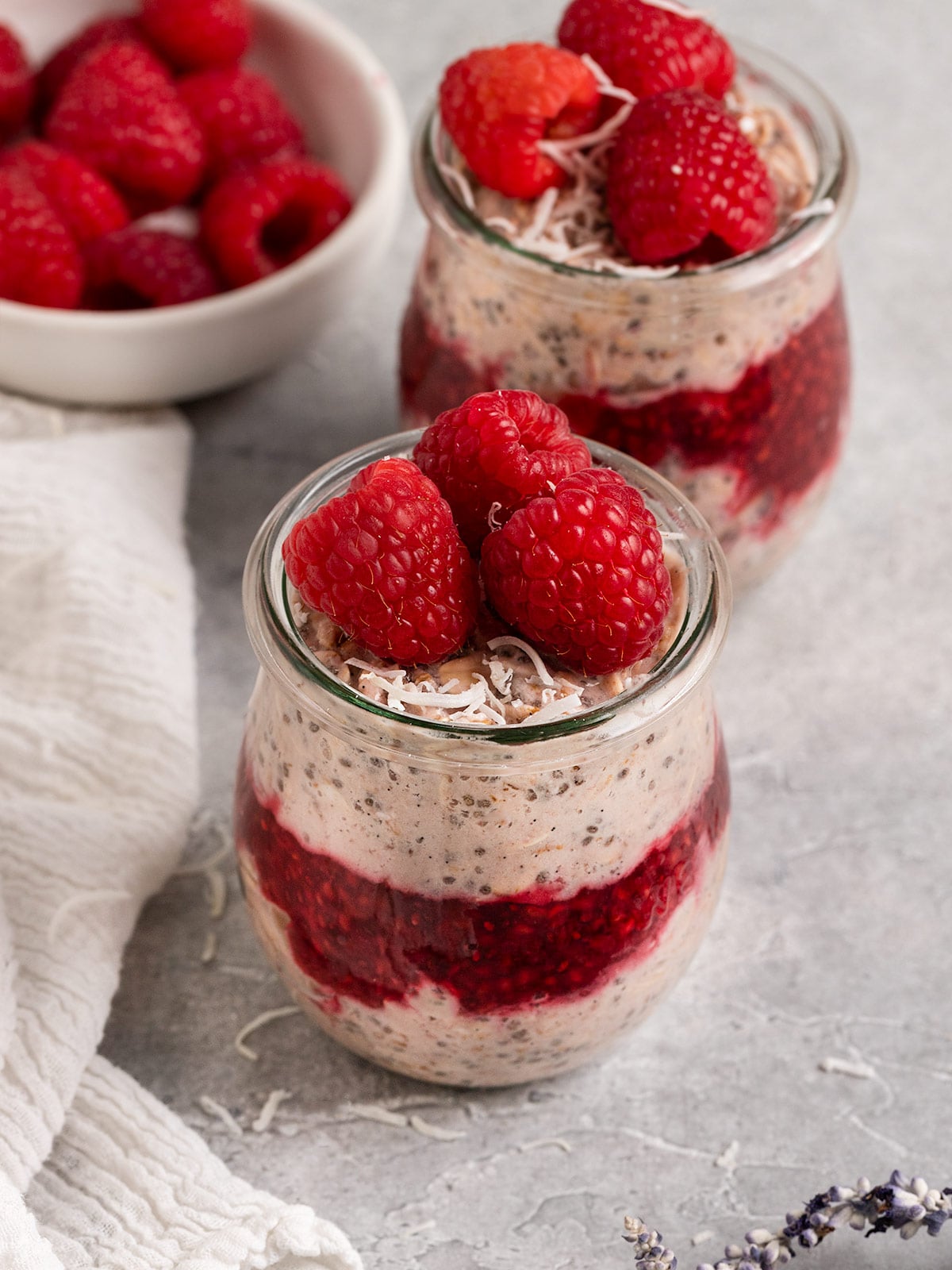 two jars of raspberry parts layered with raspberry jam and topped with fresh raspberries aside a bowl of fresh raspberries.