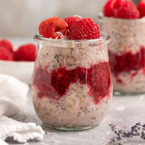 two jars of raspberry parts layered with raspberry jam and topped with fresh raspberries aside a bowl of fresh raspberries.