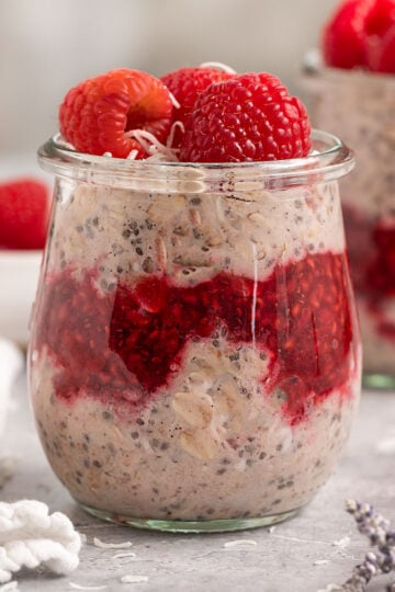 two jars of raspberry parts layered with raspberry jam and topped with fresh raspberries aside a bowl of fresh raspberries.