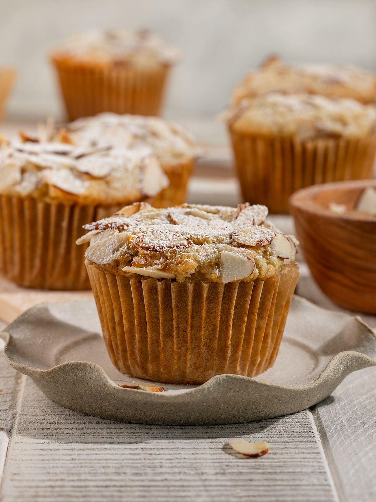 muffin topped with sliced almonds and powdered sugar on a plate with more muffins behind it.